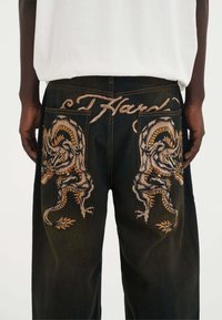 Los vaqueros de mezclilla oscura presentan diseños de dragón bordados en gris y naranja en los bolsillos traseros, con costuras decorativas arriba.