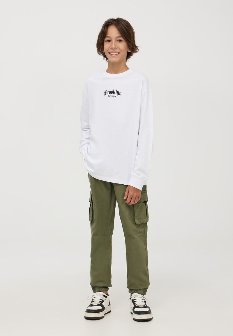 Maglietta bianca a maniche lunghe con stampa "Brooklyn", abbinata a pantaloni cargo verde oliva con tasche e scarpe da ginnastica in bianco e nero.