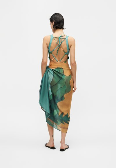 Femme portant un haut à bretelles turquoise à dos ouvert et une jupe portefeuille à motifs verts et marron, debout de dos à la caméra.