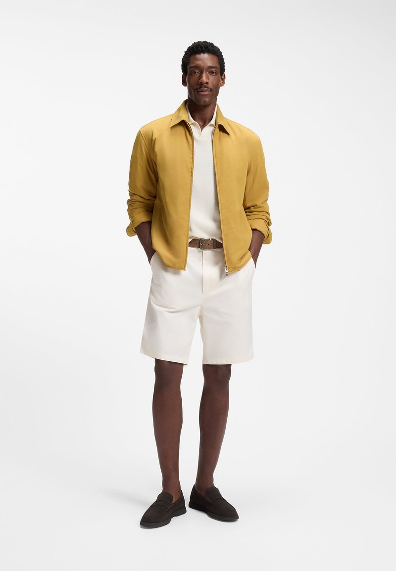 Homme debout les mains dans les poches portant une veste moutarde, une chemise blanche, un short crème, une ceinture marron et des chaussures marron foncé sur fond blanc.