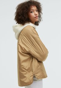 Beige Pufferjacke mit einer weichen, weißen Fleece-gefütterten Kapuze. Sie hat einen lässigen Schnitt, seitliche Taschen und einen leicht verlängerten Rücken.