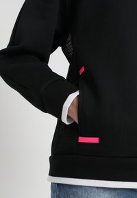 Sudadera negra con un panel de malla texturizada, que presenta detalles en rosa en los bolsillos y una capa interior blanca en los puños.