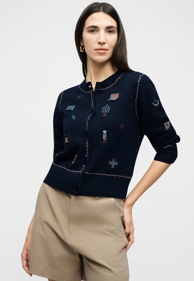 Γυναίκα με μακριά σκούρα μαλλιά, φοράει ένα μπλε-σκούρο κουμπωτό ζακέτα με πολύχρωμα κεντημένα σχέδια και μπεζ ψηλόμεσο σορτς, σε απλό φόντο.