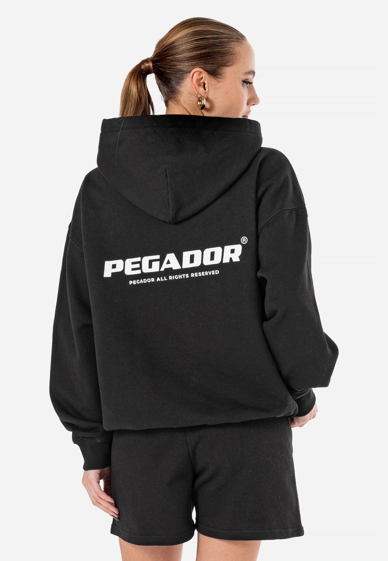 Čierna mikina s kapucňou a bielym logom "PEGADOR" na chrbte. V sprievode čiernych šortiek vyrobených z mäkkej bavlnenej tkaniny.