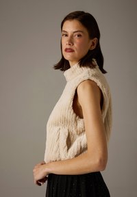 Beige, strukturierte ärmellose Weste mit hohem Kragen und Seitentaschen, aus weichem Stoff mit sichtbaren Nahtdetails, kombiniert mit einem schwarzen Rock.