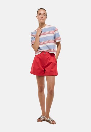 Femme portant un t-shirt rayé à manches courtes, un short rouge taille haute et des sandales blanches, debout avec une main dans la poche et l’autre près du cou.