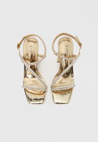 Sandale cu toc aurii, cu design cu vârf pătrat, prevăzute cu bretele strălucitoare cu strasuri și finisaj metalic. Curea reglabilă pentru gleznă inclusă.