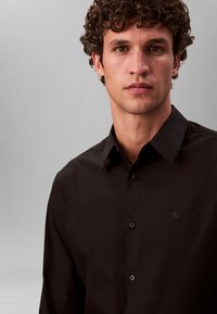 Čierna košeľa z hladkej látky s predným zapínaním na gombíky, golierom a subtílnym logom CK na ľavej strane.