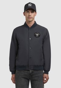 Jachetă bomber de marină cu mansete și guler elastice, închidere cu capse și o patch circular pe piept. Material texturat, design casual.