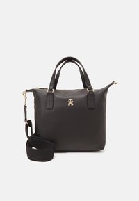 Czarna skórzana torebka na ramię z podwójnymi uchwytami, złotym logo na środku i odpinanym, szerokim czarnym paskiem na białym tle.