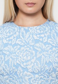 Lys blå kjole med teksturert hvit blomsterbroderi. Rund halsutringning, intrikate bladmønstre. Glatt stofffinish, ingen synlige metallbeslag.