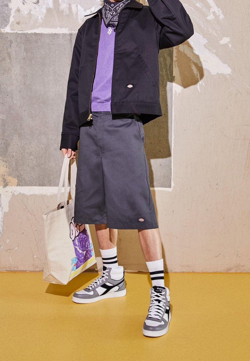 Personne portant une veste noire, une chemise violette, un short long gris, des chaussettes rayées, des baskets montantes grises, et tenant un sac fourre-tout en toile avec un imprimé coloré.