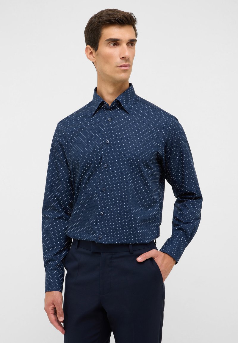 Camicia a righe blu navy con punti bianchi, maniche lunghe e colletto a punta. Presenta chiusura con bottoni sul davanti e vestibilità sagomata. Indossata con pantaloni scuri.