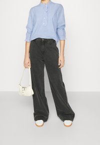 Il modello è in piedi di fronte a uno sfondo semplice, indossando una blusa azzurra con balze e pantaloni neri larghi, mentre tiene in mano una piccola borsa bianca.