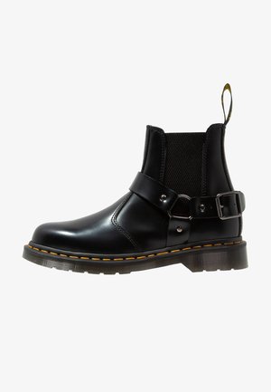 Bottine en cuir noir avec une bride à boucle latérale, un panneau élastique, une couture jaune autour de la semelle et une languette à l’arrière.