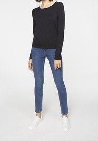 Kvinna som bär en svart långärmad texturerad topp, blå skinny jeans och vita sneakers, stående mot en enkel vit bakgrund.