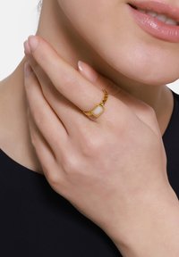 Goudkleurige ring met een gedraaid band en een rechthoekige steen setting, gedragen op een hand met neutrale nagellak tegen een donkere achtergrond.