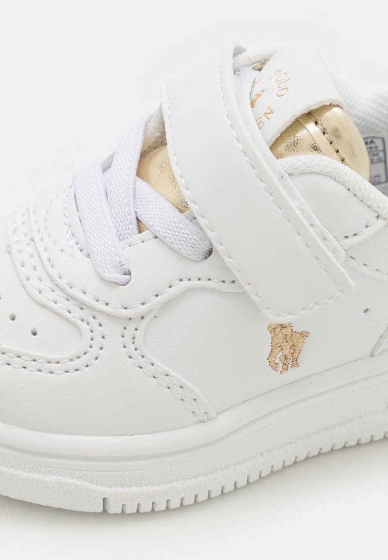 Baskets en cuir blanc avec un accent doré, une bande velcro et un logo en relief. Présente une semelle texturée et des coutures décoratives.
