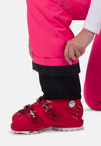 Person justerar det svarta snöbyxornas gaita över den klara röda skidstöveln med spännen och vit sula, iklädd rosa skidbyxor och vit ärm.