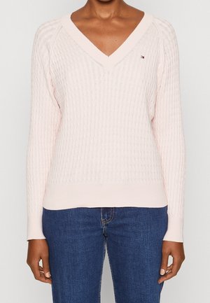 Kvinde iført en lyserød kabelstrikket V-hals sweater og blå jeans, vist fra nakke til hofter mod en ensfarvet baggrund.