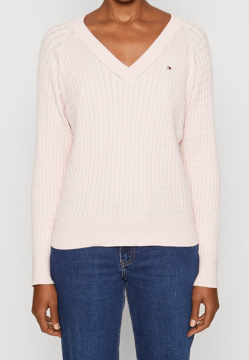 Femme portant un pull en maille torsadée rose clair à col en V et un jean bleu, visible du cou aux hanches sur un fond uni.