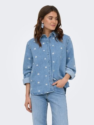 Vrouw met bruin haar die een lichtblauw denimhemd draagt met witte madeliefjesborduursel, bijpassende lichtblauwe jeans en blauwe geometrische oorbellen.