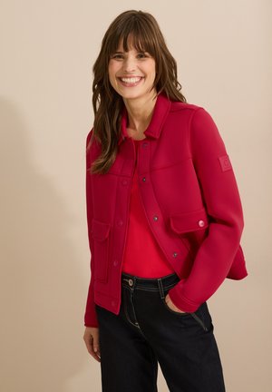 Veste softshell rouge avec un col, deux poches avant et des fermetures à pression, portée sur un haut rouge, associée à un jean foncé.