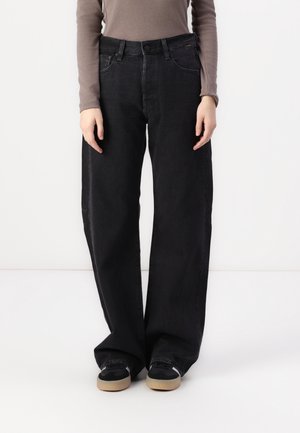 Osoba nosząca czarne szerokie jeansy, brązową bluzkę z długim rękawem i czarne trampki z beżowymi podeszwami, stoi na białym tle.