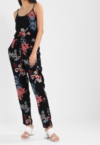 Zwart bloemen jumpsuit met rode, roze en blauwe patronen; spaghettibandjes, cinched taille en taps toelopende benen; gecombineerd met witte blokhak sandalen.