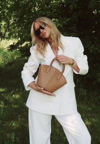 Tan leather handbag with a tapered base and two short handles. Smooth texture with visible stitching detail. Model wears a white oversized blazer.