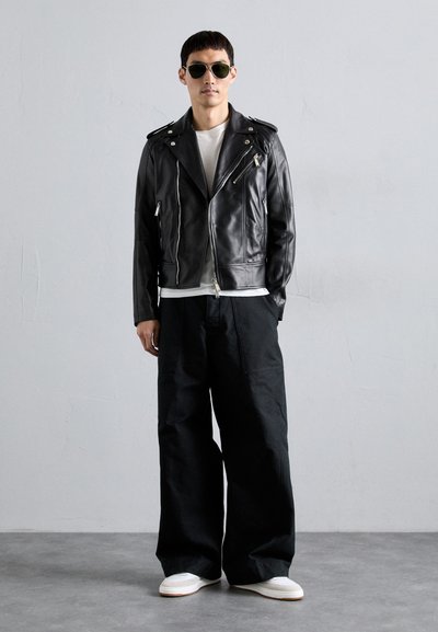 Giacca da moto in pelle nera con hardware argentato, abbinata a pantaloni neri larghi e sneakers bianche, indossata sopra una camicia bianca.