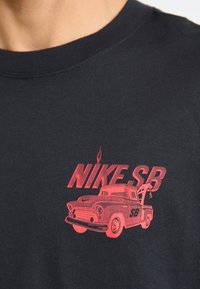 Musta puuvillane T-särk, millel on punane graafika vintage veoki kohta, millel on tekst "NIKE SB" ning detailid veoki esiosas ja tasandikul.