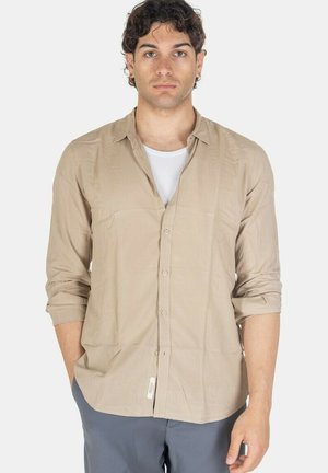 Giovane uomo con capelli scuri e ricci che indossa una camicia beige abbottonata sopra una canottiera bianca e pantaloni grigi, in piedi davanti a uno sfondo bianco.