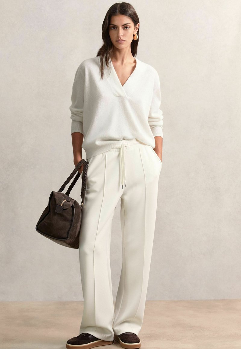 Pull blanc texturé avec un col en V. Pantalons crème taille haute avec des coutures latérales. Sac à main en daim marron et chaussures sombres avec des semelles claires.