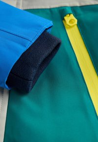 Spalvinga striukė su mėlyna rankove, tamsiai mėlynu ribbingu rankogaliu ir geltonu užtrauktuko akcentu ant žalios audinio fono.