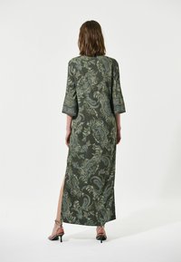 Lang groene jurk met paisleypatroon, driekwartmouwen, zijsplit en gedetailleerde zomen. Gecombineerd met schoenen met bandjes, wat een soepele textuur laat zien.