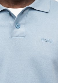 Polo en maille bleu clair avec deux boutons et "BOSS" brodé sur la poitrine, porté sur un sous-vêtement blanc.