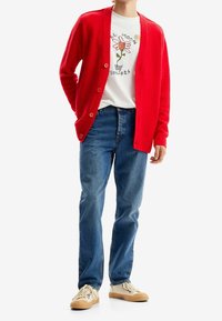 Cardigan rosso in lana con una finitura testurizzata, indossato sopra una maglietta bianca con un design di fiori ricamato, abbinato a jeans blu e sneakers beige.