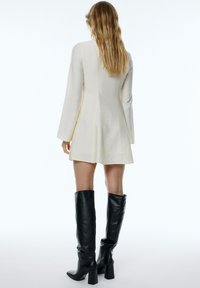 Kurzes cremefarbenes Kleid mit langen Ärmeln, strukturiertem Stoff, weit schwingendem Rock und schwarzen kniehohen Stiefeln mit hohen Absätzen. Glatte Silhouette mit kleinen Nähten.