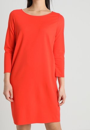 Kvinde iført en ensfarvet, knaldrød, langærmet kjole til knæet med rund halsudskæring, stående foran en lysegrå baggrund.