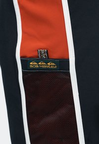 Gros plan sur un tissu avec des panneaux rouges, noirs et blancs comprenant une poche en filet et une étiquette portant l'inscription Bob Marley avec trois icônes de vagues.