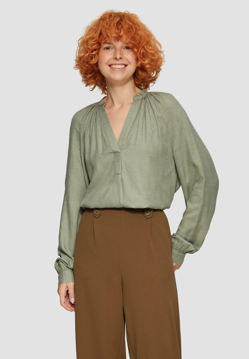 Olivgrüne Bluse mit tiefem V-Ausschnitt, gerafftem Schulterdetail und langen Ärmeln. Kombiniert mit hochgeschnittenen braunen Hosen mit Knopf-Akzenten.