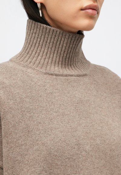 Γυναίκα που φοράει μπεζ ριγωτό πουλόβερ με ψηλό γιακά και χρυσά σκουλαρίκια σταγόνες, εμφανίζοντας το πηγούνι και το κάτω μέρος του προσώπου σε απαλό φωτισμό.