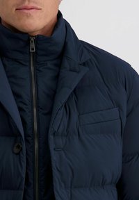 Marineblauwe pufferjack met een hoge kraag, horizontale quiltstructuur, een ritssluiting aan de voorkant en een borstzak. Inclusief een knop aan de onderzijde.