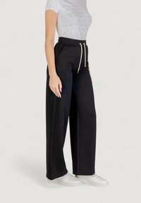 Pantalones de pierna ancha negros, confeccionados en tejido texturizado, con cintura ajustable y sutiles patrones florales. Combinados con zapatillas blancas.