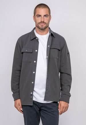 Homme aux cheveux courts et barbe, portant une chemise boutonnée gris foncé sur un t-shirt blanc et un pantalon sombre, debout devant un fond clair uni.