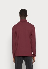 Långärmad skjorta i burgundy, mjukt tyg, med hög krage och avslappnad passform, samt sidoklipp för ökad andningsförmåga och rörelsefrihet.