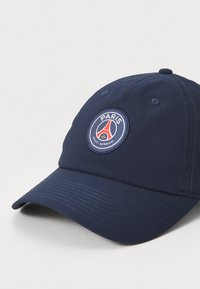 Tamsiai mėlyna audinio kepurė, su apvaliu išsiuvinėtu logotipu, kuriame baltais ir raudonais akcentais užrašytas "PARIS" ir "SAINT-GERMAIN".