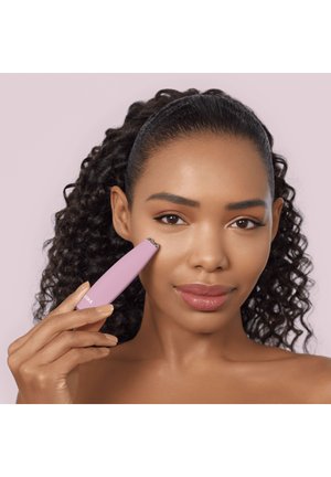 Femme aux cheveux bouclés utilisant un appareil facial rose sur sa joue devant un fond rose clair.