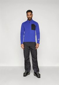 Άνδρας που στέκεται με μπλε μπουφάν με φερμουάρ, μαύρη τσέπη στο στήθος, μαύρο παντελόνι και μαύρες μπότες του σκι, μπροστά σε απλό φόντο.